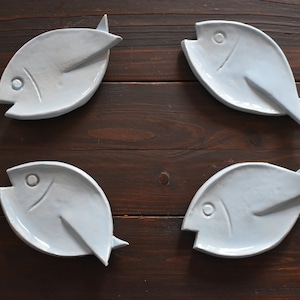 Könnte beinhalten: Vier hellblaue Keramikschalen in Fischform. Jede Schale hat eine glatte, glänzende Oberfläche mit einer einfachen eingravierten Linie, die das Auge darstellt, und einem erhabenen Flossendetail. Die fischförmigen Teller sind in einem Raster angeordnet.