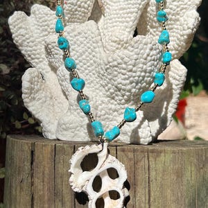 May include: A handcrafted necklace featuring turquoise beads and gold-colored accents. The necklace has a large, white seashell pendant. The necklace is displayed on a white coral-like structure, set against a natural wood background.