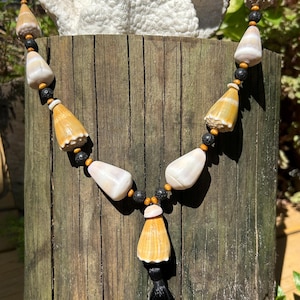 Op de afbeelding: Een handgemaakte ketting met natuurlijke elementen. De ketting heeft crèmekleurige schelpkralen, afgewisseld met zwarte en houten kralen. Een zwarte kwast hangt in het midden en voegt elegantie toe aan dit unieke sieraad.