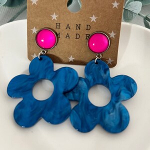 Könnte beinhalten: Ein Paar blaue und weiße, wirbelnde Blumen-Ohrhänger mit rosafarbenen, schillernden runden Oberteilen. Die Ohrringe befinden sich auf einer Karte mit der Aufschrift "HAND MADE".
