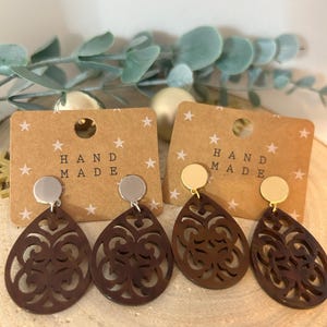 Könnte beinhalten: Paar braune Holzohrringe mit filigranen Ausschnitten. Die Ohrringe sind tropfenförmig und hängen an silbernen und goldenen Ohrhaken. Die Karte hinter den Ohrringen lautet "Hand Made".