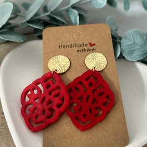 Może przedstawiać: Para czerwonych, ażurowych kolczyków ze złotymi, okrągłymi ćwiekami. Kolczyki są prezentowane na brązowej karcie z napisem "Handmade with love". Kolczyki mają geometryczny wzór.