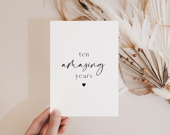 May include: A white card with the text "ten amazing years" in black script, with a small heart below. The card is held in a hand, with dried flowers in the background. The overall theme is celebration and milestone events.