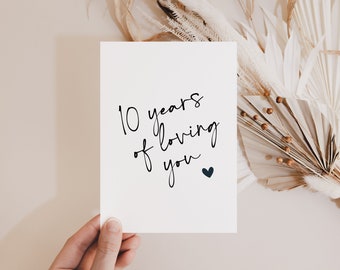 May include: A white card with the text "10 years of loving you" in black script, with a small navy blue heart. The card is held in a hand, with dried flowers in the background.