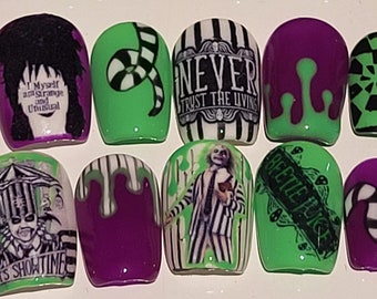 May include: Set of ten false fingernails with a green and purple base. Each nail has a different design featuring characters and quotes from the movie "Beetlejuice". Designs include "Never Trust the Living", "It's Show Time", and "Beetlejuice" written on a tombstone.
