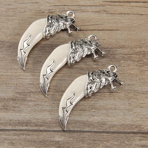 May include: Three fang-shaped pendant charms. Each charm features a cream-coloured tooth and a silver-coloured wolf head. The fangs have a decorative silver design along the edges. Each charm has a small loop at the top for attaching to a necklace or bracelet.