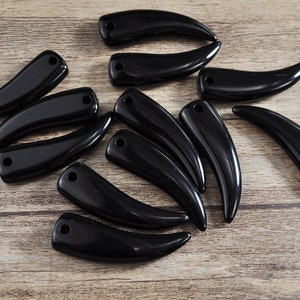 Può includere: Una collezione di dieci ciondoli neri a forma di corno con un foro nella parte superiore per la corda. I ciondoli sono realizzati in un materiale liscio e lucido e sono disposti in uno schema casuale su una superficie di legno.