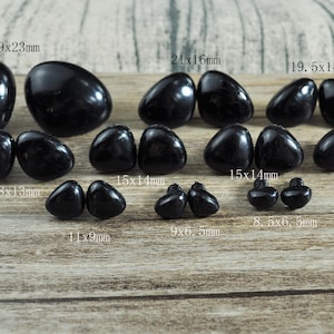 Puede incluir: Surtido de ojos de seguridad de plástico negro en varios tamaños, que van desde 8,5x6,5 mm hasta 29x23 mm. Los ojos se muestran sobre una superficie de madera.