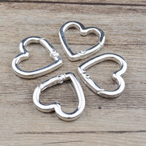 May include: Four silver-colored heart-shaped carabiner clips. Each clip has a spring-loaded gate for secure closure. The clips are arranged on a light brown wooden surface.