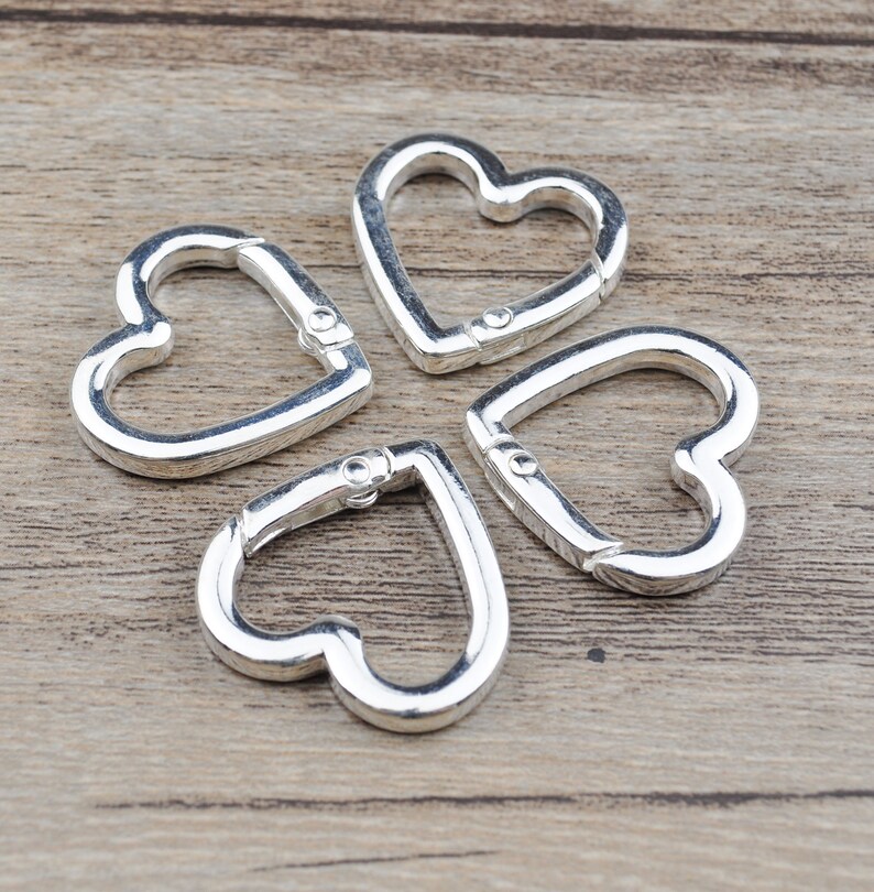 May include: Four silver-colored heart-shaped carabiner clips. Each clip has a spring-loaded gate for secure closure. The clips are arranged on a light brown wooden surface.