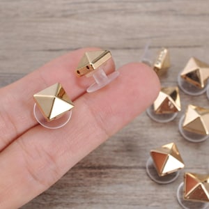 May include: A collection of gold-toned pyramid-shaped studs, each featuring a smooth, reflective surface and a clear plastic backing. These small, square-based metal components are designed for attachment to fabric or other materials for decorative purposes.