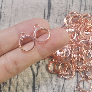 Peut inclure: Gros plan sur un tas de crochets d'oreille en or rose. Les crochets sont petits et ronds, avec une petite boucle en haut. Il y a deux crochets au premier plan, tenus par une main.