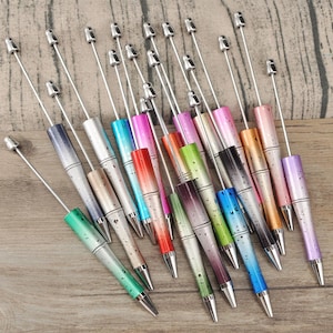 Op de afbeelding: Een verzameling kleurrijke pennen met zilveren accenten. De pennen hebben een gradiëntontwerp in verschillende kleuren, waaronder blauw, roze, groen en oranje. De pennen liggen op een houten oppervlak.