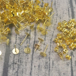 May include: A large quantity of gold-toned earring backs and earring posts. The earring backs are small and have a rounded shape. The earring posts are longer and have a flat base.