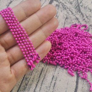 May include: A close-up of a pile of bright pink metal chain, with a few strands held in a hand. The chain is made of small, round links.