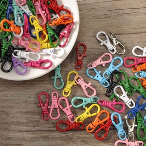 May include: A selection of brightly coloured metal swivel clasps in shades of pink, yellow, green, and blue, are scattered on a wooden surface and in a white dish. Some clasps are silver or gold-toned. These clasps are used for keychains and craft projects.