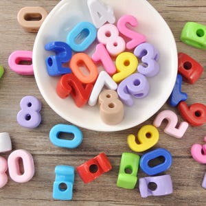 Puede incluir: Una colección de cuentas de números de plástico de colores, en varios colores, incluyendo rosa, azul, amarillo y verde. Algunas cuentas están en un cuenco blanco, mientras que otras están esparcidas sobre una superficie de madera. Los números van del 0 al 9, con algunas cuentas 30.