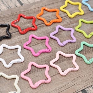 Puede incluir: Una colección de cierres metálicos en forma de estrella en varios colores, incluyendo rojo, naranja, amarillo, verde, azul, rosa, morado y marrón. Los cierres están dispuestos sobre una superficie de madera, mostrando sus vibrantes tonos y diseño.