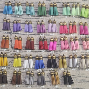 May include: Assortment of 39 colorful suede tassels with gold metal tops. The tassels are arranged in rows on a wooden surface.