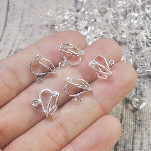 May include: Five silver-toned earring clips with a heart-shaped design. Each clip has a small clear rhinestone in the center.