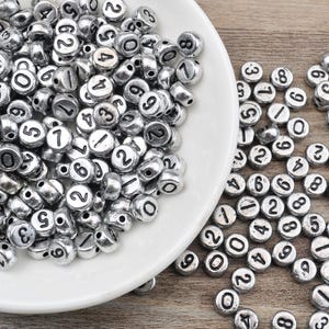 Puede incluir: Un bol lleno de cuentas de metal plateado, cada una con un número negro impreso en ella. Los números van del 0 al 9.