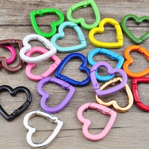 May include: A collection of heart-shaped carabiner clips in various colors, including green, blue, pink, and gold. These metal clips are designed with a spring-loaded gate for secure attachment. The clips are arranged on a light brown wooden surface.