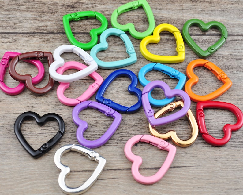May include: A collection of heart-shaped carabiner clips in various colors, including green, blue, pink, and gold. These metal clips are designed with a spring-loaded gate for secure attachment. The clips are arranged on a light brown wooden surface.