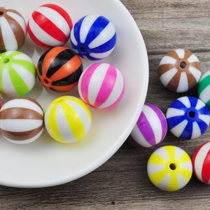 Può includere: Una collezione di perline di plastica colorate a righe e con motivi in una ciotola bianca. Le perline sono di vari colori, tra cui rosso, blu, verde, giallo, rosa e marrone.