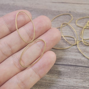 Pode incluir: Um close-up de uma mão segurando dois anéis de metal de forma ovalada de cor dourada. Os anéis são lisos e têm uma textura ligeiramente martelada. Existem vários outros anéis do mesmo estilo espalhados sobre uma superfície de madeira.