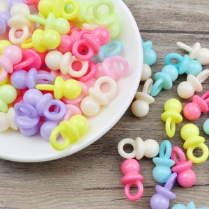 May include: A bowl filled with colorful plastic baby pacifier charms. The charms are in various pastel colors, including pink, blue, yellow, and green. The charms are arranged in a random pattern in the bowl.