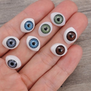 May include: A collection of eight oval-shaped craft eyes with white bases and realistic irises in various colors including blue, green, and brown. Each eye features a dark central pupil and detailed radial iris patterns, designed for use in dolls or toys.