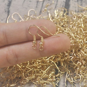 May include: A close-up of a hand holding two gold earring hooks. The hooks have a small ball at the end and a spring-loaded closure. The background is a pile of similar earring hooks.