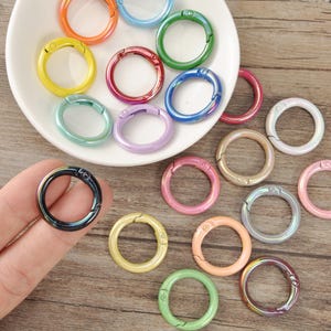 May include: A selection of colourful, round metal spring snap clasps in various iridescent shades, including blue, green, pink, and yellow. Some clasps are on a white plate, others are on a wooden surface.