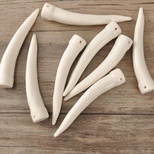 May include: A collection of off-white, curved, horn-shaped beads with a glossy finish. Each bead has a hole drilled at one end. The beads are arranged on a wooden surface.