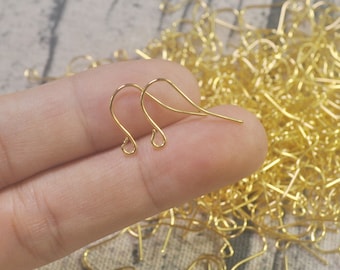May include: Two gold-colored earring hooks, also known as ear wires, are held in a hand. The hooks are made of thin metal and have a simple, classic design. The hooks are commonly used for making earrings.