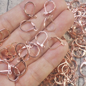 May include: A close-up of a hand holding a large quantity of rose gold earring hooks. The hooks are made of metal and have a smooth finish.
