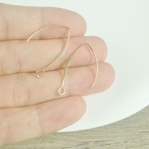 May include: Two gold-toned earring hooks, made of thin wire, are shown on a hand. The hooks are curved and have a small loop at the end.