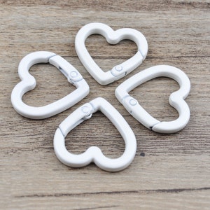 May include: Four white heart-shaped carabiner clips arranged on a wooden surface. Each clip has a hinged opening mechanism. The clips are made of a solid, opaque white material.