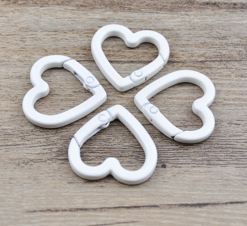 May include: Four white heart-shaped carabiner clips arranged on a wooden surface. Each clip has a hinged opening mechanism. The clips are made of a solid, opaque white material.