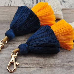May include: Two keychains with blue, yellow, and white tassel charms. The tassels are attached to gold metal key rings.