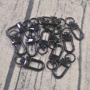 Pode incluir: Um grupo de chaveiros de metal preto com fecho giratório. Os chaveiros estão dispostos em uma pilha sobre uma superfície de tecido texturizada.