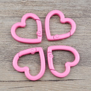 May include: Four pink heart-shaped carabiner clips. Each clip has a hinged opening with a small metal clasp. The clips are made of a smooth, solid material and are arranged on a light brown wooden surface.