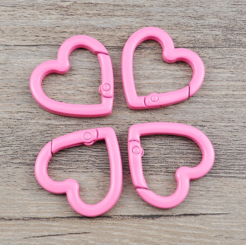 May include: Four pink heart-shaped carabiner clips. Each clip has a hinged opening with a small metal clasp. The clips are made of a smooth, solid material and are arranged on a light brown wooden surface.