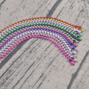 May include: A rainbow assortment of colorful metal chain links, each with a different color, arranged in a semi-circle on a textured fabric background.