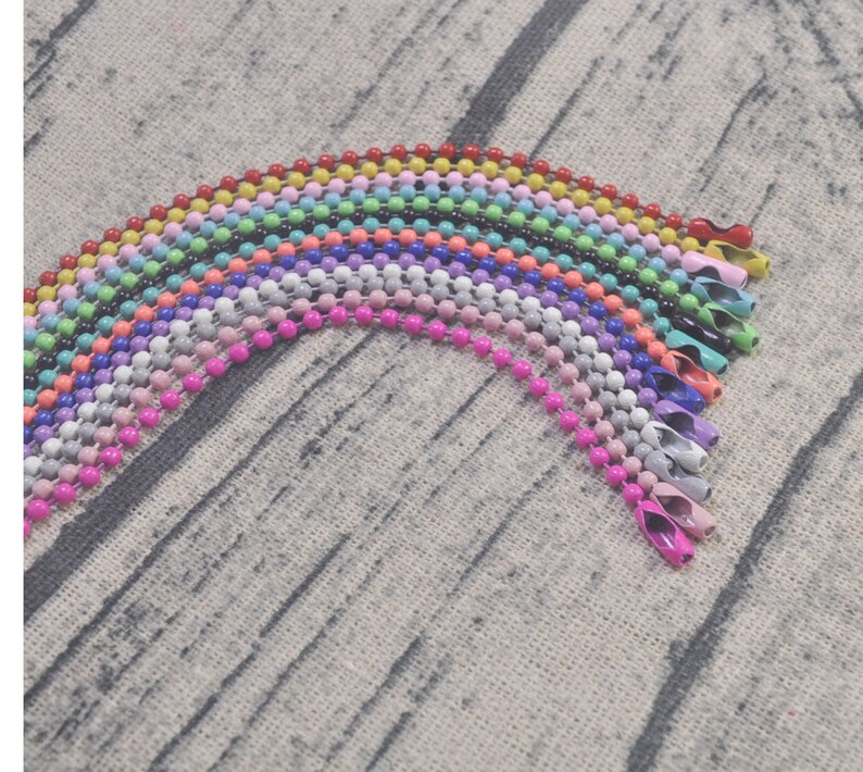 May include: A rainbow assortment of colorful metal chain links, each with a different color, arranged in a semi-circle on a textured fabric background.