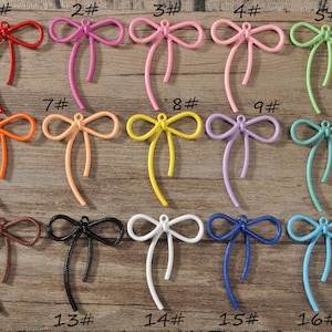 Peut inclure: Une collection de seize breloques en forme de nœuds colorés, dans diverses teintes, dont rouge, rose, vert, orange, jaune, violet, marron, noir, blanc et bleu. Chaque breloque a une boucle en haut et une longue queue fine et texturée. Les breloques sont disposées sur une surface en bois.