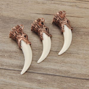 May include: Three bronze-colored wolf head pendants with large, curved, off-white fangs. The pendants are detailed with textured fur and facial features, and have a small loop at the top for hanging. The background is a light brown wooden surface.