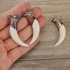 Peut inclure: Trois breloques en forme de dents d'animaux. Chaque breloque a une dent blanche et incurvée et une tête d'animal métallique aux couleurs de l'arc-en-ciel avec une boucle pour suspendre. Les breloques sont présentées sur une surface en bois.