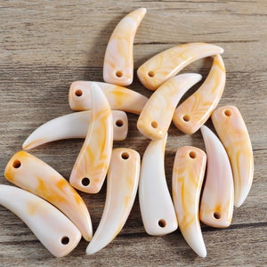 Può includere: Una collezione di 12 ciondoli a forma di dente di conchiglia bianca e arancione con un foro praticato nella parte superiore per la corda. I ciondoli hanno la forma di un dente e hanno un aspetto naturale e organico.