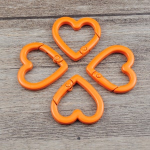 May include: Four orange heart-shaped carabiner clips arranged in a cloverleaf pattern. Each clip has a spring-loaded gate for secure attachment. The clips are made of a smooth, solid material and are set against a wooden background.